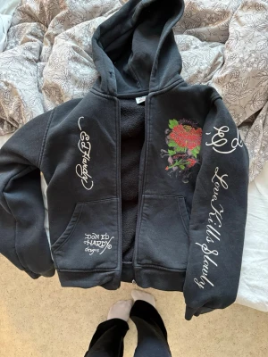 Svart Vintage Ed Hardy hoodie - Svart hoodie från Ed Hardy med dragkedja, stor huva och fickor fram. Snygga vita tryck på ärmar och bröst, bland annat texten 'Love Kills Slowly' och ett färgglatt hjärta med rosor på bröstet. Mjuk insida och klassisk streetwear-vibe.