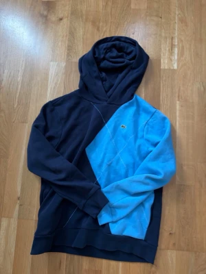 Blå och marinblå hoodie från Lacoste - Säljer en snygg hoodie från Lacoste i tvåfärgad design med marinblå och ljusblå paneler. Hoodien har en klassisk huva och den ikoniska krokodilloggan på bröstet. Tillverkad i mjukt bomullsmaterial för skön känsla. Perfekt för en avslappnad streetwear-look.