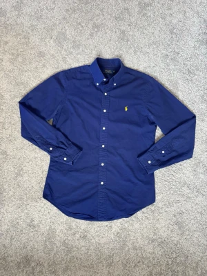 Marinblå skjorta från Polo Ralph Lauren - Klassisk marinblå skjorta från Polo Ralph Lauren i slim fit. Skjortan har långa ärmar, knappar framtill och en gul broderad logga på bröstet. Tillverkad i bomull och har button-down krage för en stilren look.