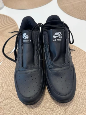 Nike Air Force 1 helsvarta sneakers - Säljer ett par klassiska Nike Air Force 1 sneakers i helsvart läder. Skorna har perforerad tå, svart sula och ikonisk Nike Swoosh på sidan. Tjock plös med vit Nike Air-logga. Perfekta för dig som gillar stilrena och tidlösa sneakers.