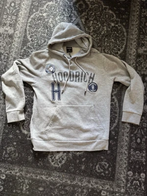 Hoodrich Hoodie - Säljer en grå hoodie från HOODRICH med huva och känguruficka. Framsidan har stora tryck och broderade detaljer i blått och vitt, inklusive HOODRICH-loggan och flera patches. Klassisk passform och mjukt material, perfekt för en avslappnad streetwear-look. Storlek M och är mycket bra skick! Hör av dig vid frågor eller liknande!