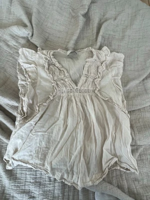 Beige volangblus från Neo Noir - Supersöt beige blus från Neo Noir med volangdetaljer och djup v-ringning. Blusen har fina broderade partier och är tillverkad i ett tunt, luftigt tyg som ger en romantisk vibe. Perfekt till jeans eller kjol för en trendig look.