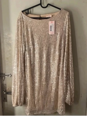 Glittrig kortklänning - Supersnygg kort klänning i ljus champagnefärg med massor av paljetter som ger ett riktigt glittrigt intryck. Klänningen har långa ärmar och en öppen ryggdetalj. Perfekt för dig som vill sticka ut och glänsa. Helt ny oanvänd med prislapp kvar!