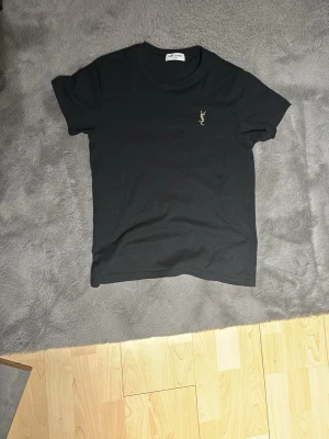 Svart t-shirt från Saint Laurent - Snygg svart t-shirt med guldfärgad Saint Laurent -logga broderad på bröstet. Klassisk rund halsringning och korta ärmar. Perfekt basplagg som passar till det mesta och är enkel att styla. Tillverkad i mjuk bomull för skön känsla hela dagen. Hör av er vid minsta fundering 💫