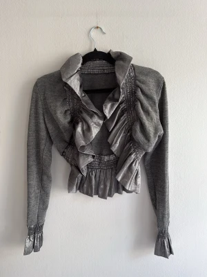 Grå volangkofta med satindetaljer - Snygg grå kofta med långa ärmar och volangkant i glansigt satinmaterial längs krage, midja och ärmslut. Kortare modell med smockdetaljer som ger en unik och trendig look. Perfekt för dig som vill sticka ut med din outfit.