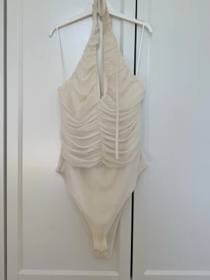 Krämvit halterneck body - Säljer en krämvit body med halterneck och djup urringning. Body har rynkade detaljer framtill och knytband i nacken. Materialet ser ut att vara stretchigt och följsamt.