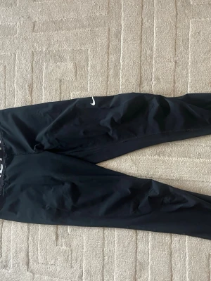 Svarta Nike Pro leggings - Säljer ett par svarta Nike Pro leggings med vit logga på benet och bred resår i midjan med Nike Pro-tryck. Tight passform och stretchigt material som passar perfekt till träning eller chill. Klassisk sportig look.