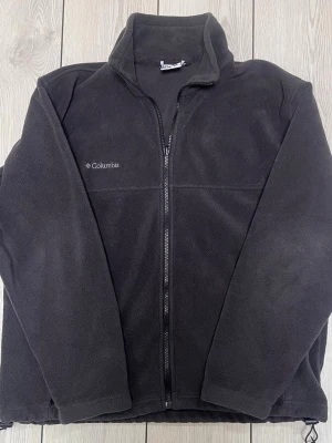 Svart fleecejacka från Columbia - Mysig svart fleecejacka från ikonsika märket Columbia. Jackan har diskret logga broderad på bröstet och justerbar nederkant med dragsko. Perfekt inför hlsten som kommer!