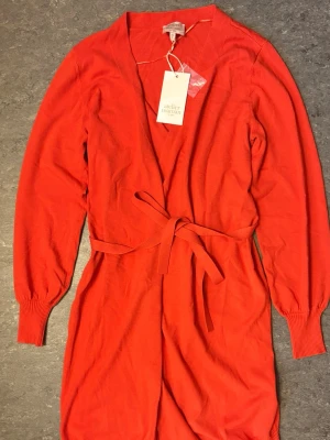 Röd jacka med knytband - Snygg röd jumpsuit från Atelier Magasin med långa ärmar och knytband i midjan. Mjuk och stretchig kvalitet, ribbade muddar vid ärmslut och benslut. Djup v-ringning framtill ger en trendig look. Perfekt för dig som vill sticka ut.