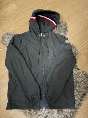 Grå Moncler Vindjacka - Snygg svart vindjacka med huva från moncler, hel dragkedja framtill och elastiska muddar. Jackan har en röd, vit och blå rand på huvan samt en patch på ärmen. Tillverkad i lätt och vindtätt material, perfekt för blåsiga dagar. Endast använd 4-5 gånger, inga tecken på användning se bilder. Storlek 2/M