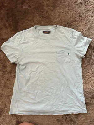 Ljusblå T-shirt med ficka - En enkel ljusblå t-shirt i bomull med rund hals och korta ärmar. På bröstet finns en liten ficka med en grön broderad detalj. Perfekt basplagg för alla tillfällen och lätt att matcha med andra kläder.