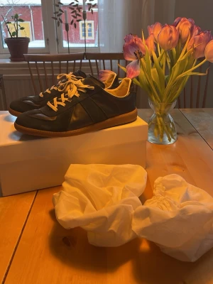 Maison Margielas blå - Box och påse följer med. Stilrena blå sneakers med vita snören och beige innersida. Skorna har en klassisk låg siluett och ovandel i både mocka och läder, samt en brun gummisula. Perfekta för dig som gillar en clean och tidlös look. En skada vid hälen (kolla bild)