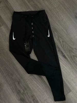 Nike aeroswift (junior-S/M) - Svarta träningsbyxor från Nike med vita loggor på båda sidor framtill. Byxorna har dragsko i midjan och är tillverkade i ett lätt och snabbtorkande material, perfekt för sport och träning. Smal passform och ribbade detaljer vid benslut. Passform S/M (160-175 ish cm) använd fåtal gånger men inte mycket synlighet på användning  