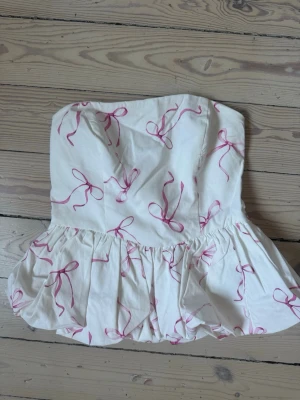 Vit tubtopp med rosa rosett-mönster - Supersöt vit tubtopp med volangkant och rosa rosetter som mönster. Toppen är axelbandslös och har en lätt puffig form nedtill. Perfekt för sommarens alla tillfällen och ger en lekfull vibe.