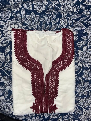 Vit kaftan med vinröd brodyr - Säljer en vit kaftan med detaljerad vinröd brodyr längs halsringningen och framtill. Plagget har en traditionell design med vackra mönster och är tillverkad i ett mjukt tyg som känns bekvämt mot huden. Perfekt för dig som gillar unika och stilrena plagg.