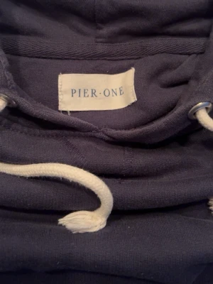 Mörkblå hoodie från Pier One - En stilren mörkblå hoodie från Pier One med klassisk huva och vita dragsnören. Hoodien har en känguruficka framtill och ribbade muddar vid ärmslut och nederkant. Perfekt för en avslappnad och casual look.