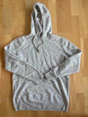Ljusgrå Kashmir Hoodie - Säljer en stilren ljusgrå hoodie med huva och dragsko. Hoodien har långa ärmar, ribbade muddar och är tillverkad i ett mjukt, stickat material som känns riktigt skönt mot huden. Perfekt för dig som gillar en enkel och clean look.