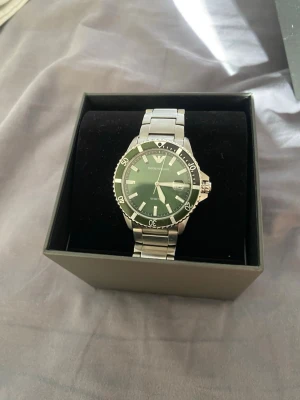 Emporio Armani silverfärgad klocka - Stilren klocka från Emporio Armani med grön urtavla och silverfärgad länk i metall. Klockan har tydliga index, datumvisning och vridbar bezel. Perfekt accessoar för dig som vill ha en klassisk men modern look. Kvitto ingår