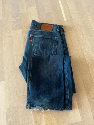 Mörkblå raka jeans med röd detalj - Snygga mörkblå jeans med rak passform och klassisk femficksdesign. Jeansen har en röd etikett på bakfickan och läderpatch i midjan. Materialet är kraftig denim som ger en tidlös look. Perfekta till sneakers eller boots. Lite slitningar nere vid fötterna!