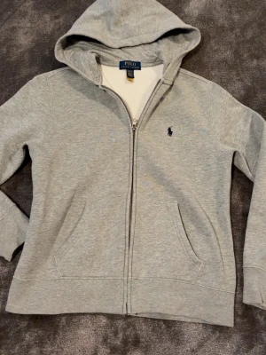 Grå zip hoodie från Polo Ralph Lauren - Snygg grå hoodie med dragkedja från Polo Ralph Lauren. Klassisk design med huva, två fickor framtill och den ikoniska lilla loggan broderad på bröstet. Tillverkad i mjukt sweatshirtmaterial som är perfekt för chill dagar.