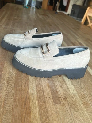 Dasia skor - Snygga beige loafers i mocka med chunky svart sula och guldfärgad kedjedetalj över foten. Skorna har en modern, fyrkantig tå och markerade sömmar som ger en trendig look. Perfekta för dig som vill ha en stilren men edgy sko till vardagslooken. Kommer med original lådan.