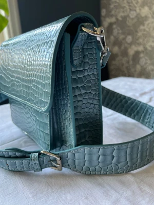 HVISK: Turkos  axelväska med krokopräglat mönster  - Snygg turkos axelväska i konstläder med krokopräglat mönster. Väskan har justerbar axelrem med silverfärgade detaljer och en stilren, boxig form. Perfekt för dig som vill ha en trendig accessoar med cool struktur. 20x17 cm