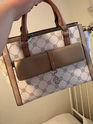 Beige och brun handväska med mönster - Snygg handväska i beige och brunt med grafiskt mönster och bruna detaljer. Väskan har två handtag, en stor ficka framtill och ett dekorativt hänge. Tillverkad i konstläder med metallspännen och dragkedja. Perfekt för dig som gillar stilrena accessoarer.köpt för 400 säljes för 100kr 