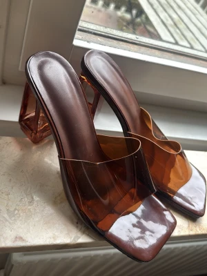 Bruna transparenta pumps med klack - Säljer ett par trendiga pumps i brun transparent plast med fyrkantig tå och bred, stabil klack. Skorna har en modern design med genomskinliga detaljer och slip-in modell. Perfekta för dig som vill sticka ut med en unik look.