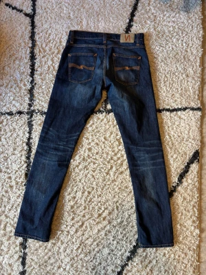 Mörkblå Nudies - Säljer ett par mörkblå Nudie jeans med slim fit och snygga slitningar. Perfekta för dig som gillar en enkel men stilren look. Varsamt använda och i gott skick.