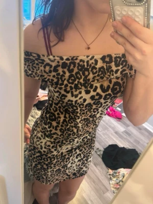 Leopardmönstrad offshoulder klänning - Säljer en kort, tight klänning med leopardmönster. Klänningen är offshoulder och har en volangdetalj upptill. Färgerna går i beige, svart och brunt. Perfekt för dig som vill sticka ut med en trendig look.