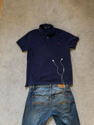 Marinblå pikétröja från Ralph Lauren - Klassisk marinblå pikétröja från Ralph Lauren med röd broderad logga på bröstet. Tröjan har krage, knappar vid halsen och är kortärmad. Perfekt för en clean och stilren look. Storlek = L.     Hör av er vid frågor! 