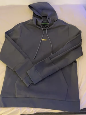 Mörkblå hoodie från BOSS - Snygg mörkblå hoodie från BOSS med klassisk huva och dragsnören. Diskret BOSS-logga på bröstet i vitt. Hoodien har en stor magficka och ribbade muddar vid ärmar och nederkant. Perfekt för en avslappnad och stilren look.
