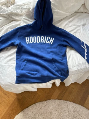 HOODRICH - Snygg blå hoodie med vit HOODRICH-text på bröstet och coolt tryck längs ena ärmen. Klassisk känguruficka framtill och stor huva. Perfekt för dig som gillar streetwear och vill sticka ut lite extra.