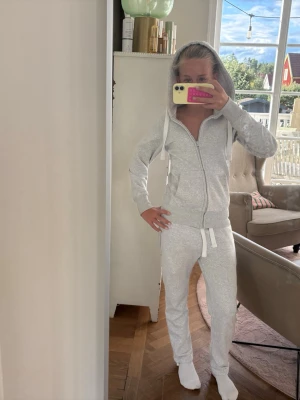 Russemerch - Supermysig ljusgrå jumpsuit i mjukt sweatshirtmaterial. Har dragkedja framtill, huva med snören och ribbade muddar vid ärmar och benslut. Perfekt för chill dagar hemma eller när du vill vara extra bekväm.