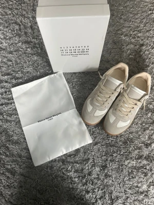 Maison Margiela vita sneakers - Snygga vita sneakers från Maison Margiela med beige detaljer och klassisk gummisula. Skorna har en låg silhuett, ovandel i läder och mocka samt vita skosnören. Perfekta för dig som gillar minimalistisk och stilren design.