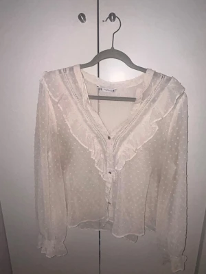 Vit blus med volanger och prickar - Säljer en romantisk vit blus med volangdetaljer längs ringningen och knäppning framtill. Blusen har ett diskret prickmönster och är tillverkad i ett tunt, lätt material. Långa ärmar med resår och volang vid ärmslut ger en extra söt touch.