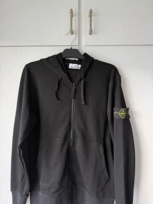 Svart hoodie från Stone Island - Säljer en svart hoodie från Stone Island med dragkedja framtill och klassisk logopatch på vänster ärm. Hoodien har huva med dragsko och två fickor framtill. Tillverkad i mjukt bomullsmaterial för en skön och avslappnad look.   (Obs, R3plika)