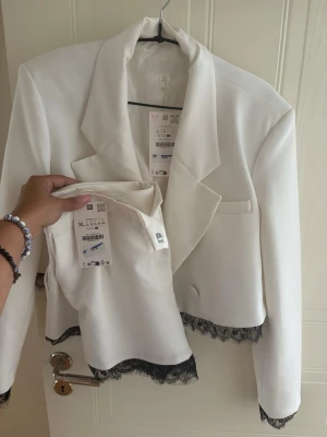 Vit croppad topp med spetskant - Snygg vit croppad topp med svart spetskant nertill. Toppen har en elegant och stilren look med raka linjer och ärmlös design. Perfekt att matcha med kavaj eller för en trendig lager-på-lager-stil.