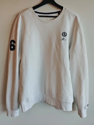 Vit sweatshirt från Henri Lloyd - Klassisk vit sweatshirt från Henri Lloyd med rund halsringning och broderad logga på bröstet. Tröjan har en svart siffra '6' på ena ärmen och är tillverkad i 90% bomull och 10% polyester för en mjuk och bekväm känsla.