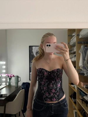 Svart body med rosa blommor - Säljer en svart body med rosa och lila blommönster. Bodyn har hjärtformad urringning och kan bäras både med och utan axelband. Materialet är glansigt och ger en elegant look. Perfekt för dig som vill ha något extra till din outfit.