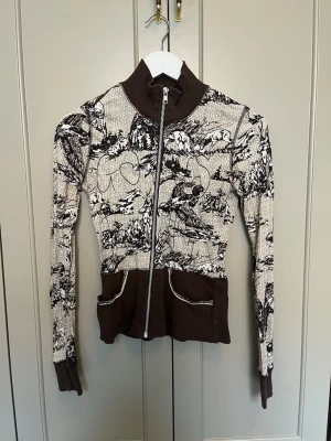 Y2k zip up från Gsus Sindustries - Unik ziptröja med naturmotiv och bruna detaljer. Tröjan har lite hög krage, långa ärmar och två fickor framtill med kontrastsömmar. Materialet är stretchigt och tunt, perfekt för dig som gillar statementplagg.