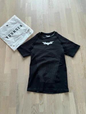 Batman Dark Knight t-shirt YoungLA - Snygg svart Batman t-shirt från YoungLA inspirerad av The Dark Knight-trilogin. Tröjan har korta ärmar, batman-logga på bröstet och coola detaljer i texturerat material. Perfekt för cosplay eller maskerad. Tillverkad i polyester och elastan för skön stretch.