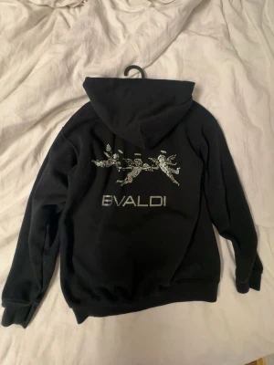 Bvaldi hoodie - Svart hoodie från Bvaldi med huva och ribbade muddar. På ryggen finns ett tryck med tre änglar och texten 'BVALDI' i glittriga stenar. Endast testad. Nypris 1100kr. Pris ej hugget i sten.