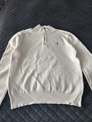 Vit stickad polo Quarter zip - Vit stickad tröja från Ralph Lauren med halv dragkedja och broderad logga på bröstet. Tröjan har ribbade muddar och krage, perfekt för en stilren och klassisk look.