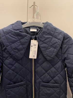 Mörkblå quiltad jacka med krage - Snygg mörkblå quiltad jacka med stor krage och dragkedja framtill. Jackan har två stora fickor och ett rutmönstrat tyg som ger en trendig look. Perfekt för kyliga dagar och enkel att matcha med olika outfits.