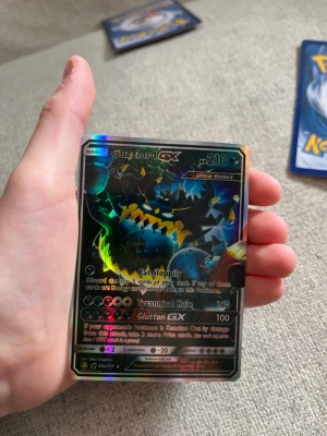 Pokémonkort Guzzlord GX Ultra Beast - Säljer ett Pokémonkort med motivet Guzzlord GX Ultra Beast. Kortet har en mörk, holografisk design med färger som svart, blått och gult. Text och attacker som Eat Sloppily, Tyrannical Hole och Glutton GX syns tydligt. Perfekt för samlare eller Pokémonfans.