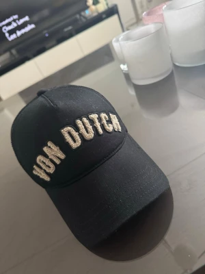 Svart Von Dutch keps - Säljer en svart keps från Von Dutch med broderad logga i vitt framtill. Klassisk böjd skärm och mesh på baksidan för extra ventilation. Justerbar passform med snapback-spänne. Perfekt för en streetwear-look.