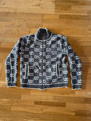 Grå Gucci cardigan tröja - Snygg grå gucci gardigan med vitt GG-mönster över hela plagget. Tröjan har hög krage, hel dragkedja framtill och två fickor. Ribbstickade muddar vid ärmslut, nederkant och krage ger en schysst passform. Perfekt för dig som gillar streetstyle och loggor.
