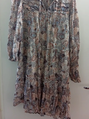 Mönstrad långklänning från Part Two - Långklänning från Part Two med bohemiskt blommigt mönster i beige, brun och blå. Klänningen har långa ärmar, v-ringning och volangdetaljer. Tillverkad i ett lätt och luftigt material, perfekt för varmare dagar.