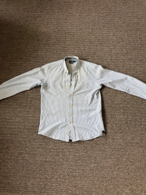Randig skjorta med knappar - Snygg vit skjorta med tunna blå ränder och klassisk krage. Skjortan har långa ärmar, knappar framtill och på manschetterna samt en liten broderad logga på bröstet. Perfekt till jeans eller chinos för en clean look.